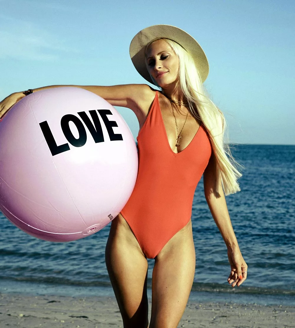 Une femme cougar en maillot de bain au bord de la plage porte un gros ballon rose sous le bras sur lequel est écris : LOVE. Elle sourit car elle est heureuse